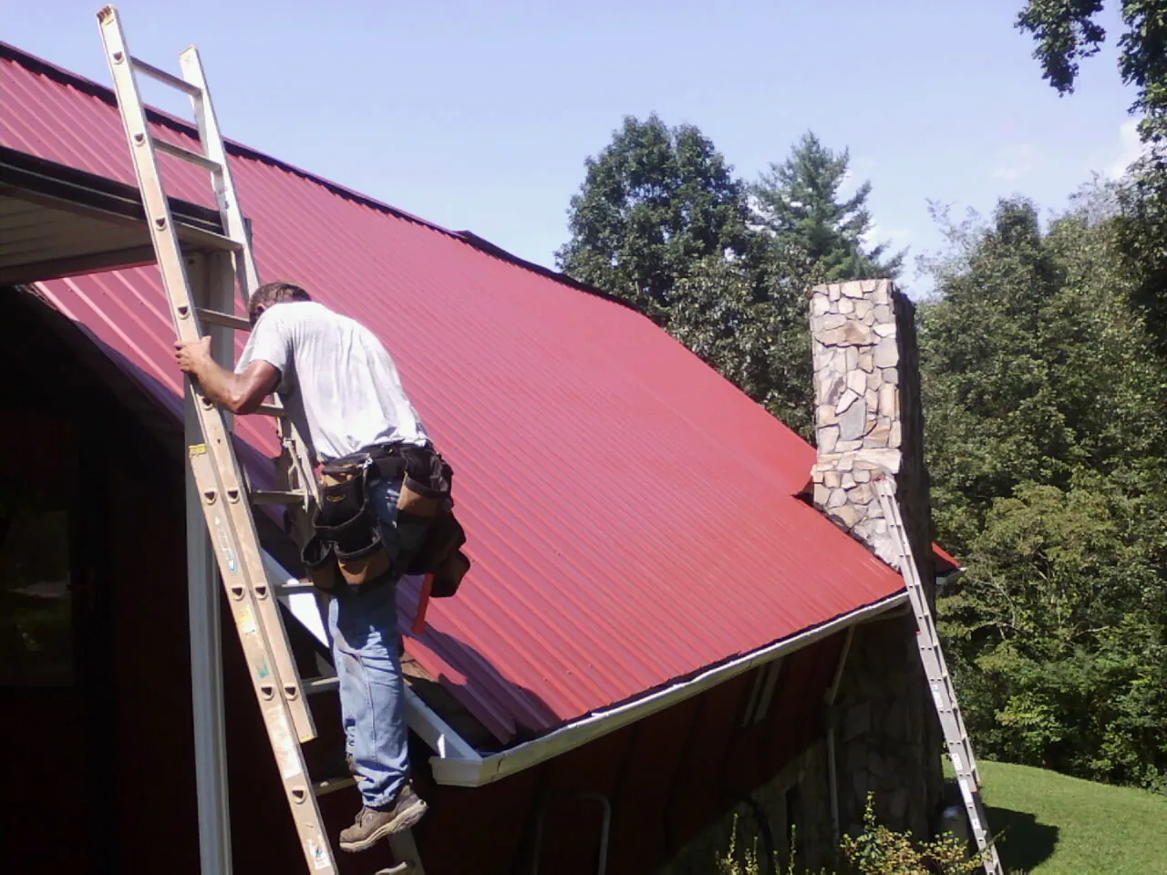 Roofer on a ladder beside a red metal roof and stone chimney — Soffit & Fascia Repair by Roofing Brothers in Scappoose